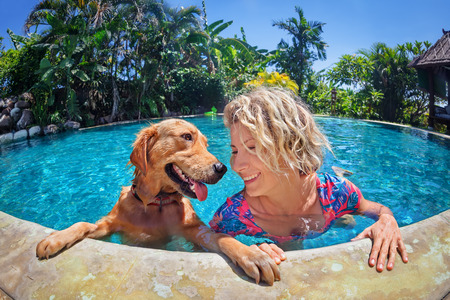 funny portrait of smiley woman playing with fun and training golden retriever puppy in outdoor swimming pool. popular dog like companion, outdoor activity and game with family pet on summer holiday